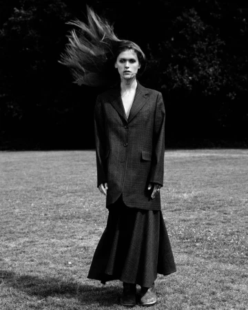 Monochrome portrait of a woman wearing a blazer and skirt, standing in a grassy field, hair blowing in the wind, against a dark background.