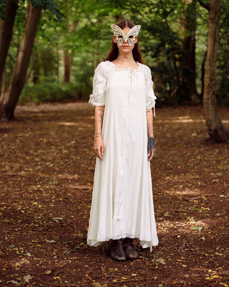 A woman wearing a long white dress and a butterfly mask stands in a forest, illuminated by soft, natural light filtering through the trees.