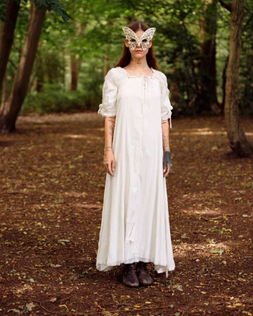 A woman wearing a long white dress and a butterfly mask stands in a forest, illuminated by soft, natural light filtering through the trees.