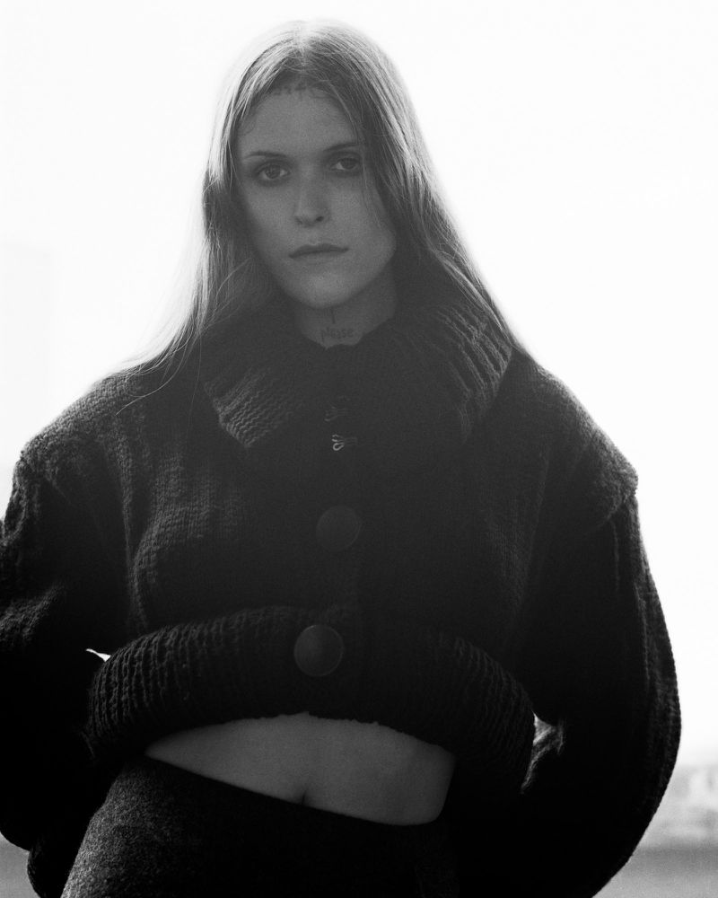 Ethel Cain posing in a black cropped chunky knit cardigan against a bright backlit background in a black and white portrait.