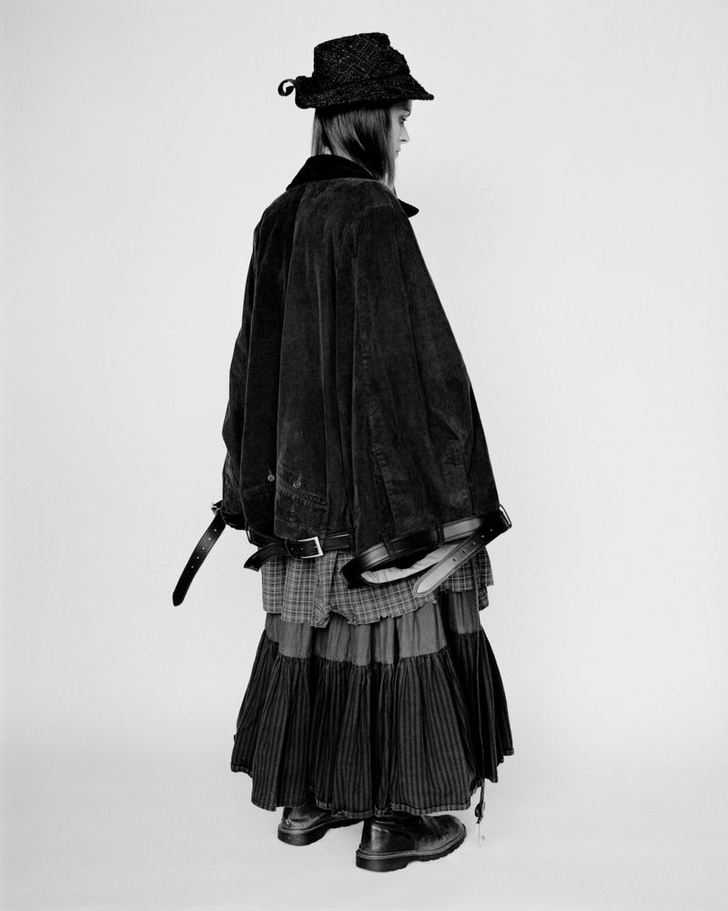 Black and white fashion portrait showcases an individual wearing a hat, cape, skirt, and boots standing against a clean, neutral backdrop.