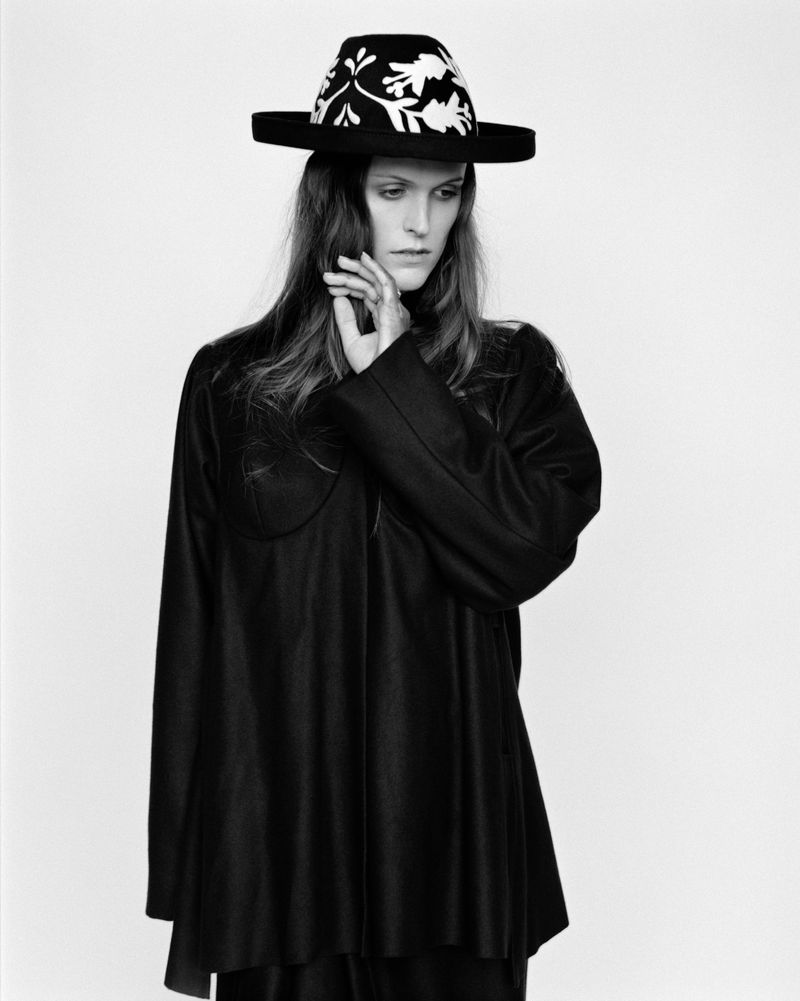 Monochromatic fashion portrait of a model wearing a black hat with white floral accents and long tunic in a minimalist photo studio.