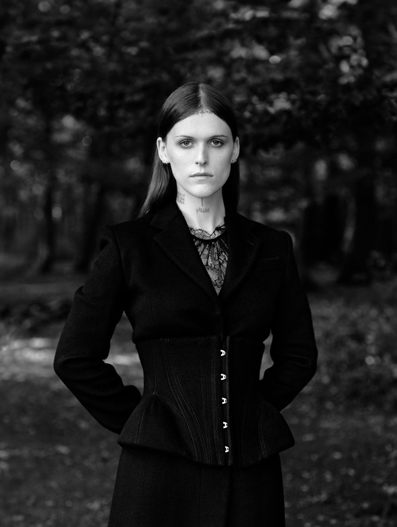 Portrait of a woman in black jacket and corset standing in a forest with dark lighting, monochrome colour palette.