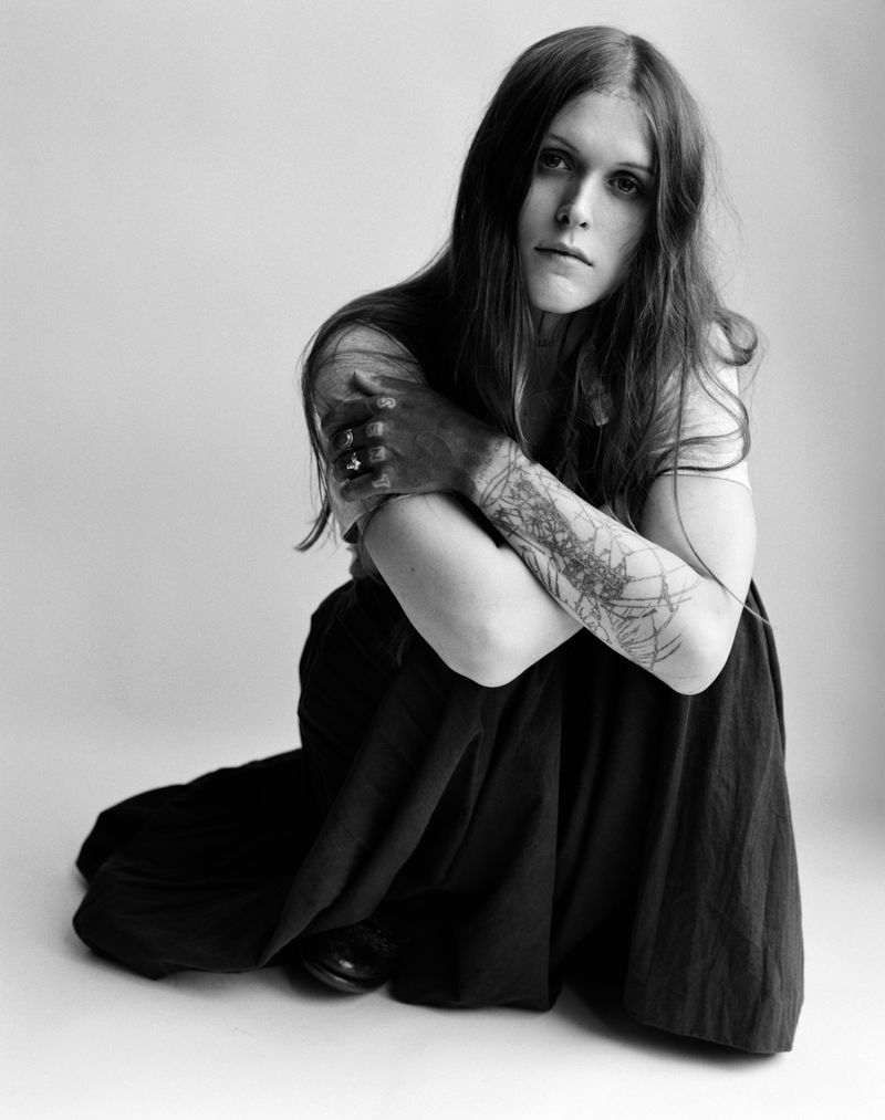 Monochrome portrait of a woman with long hair, seated with arms crossed, tattoo visible on arm, gazing directly at the camera.