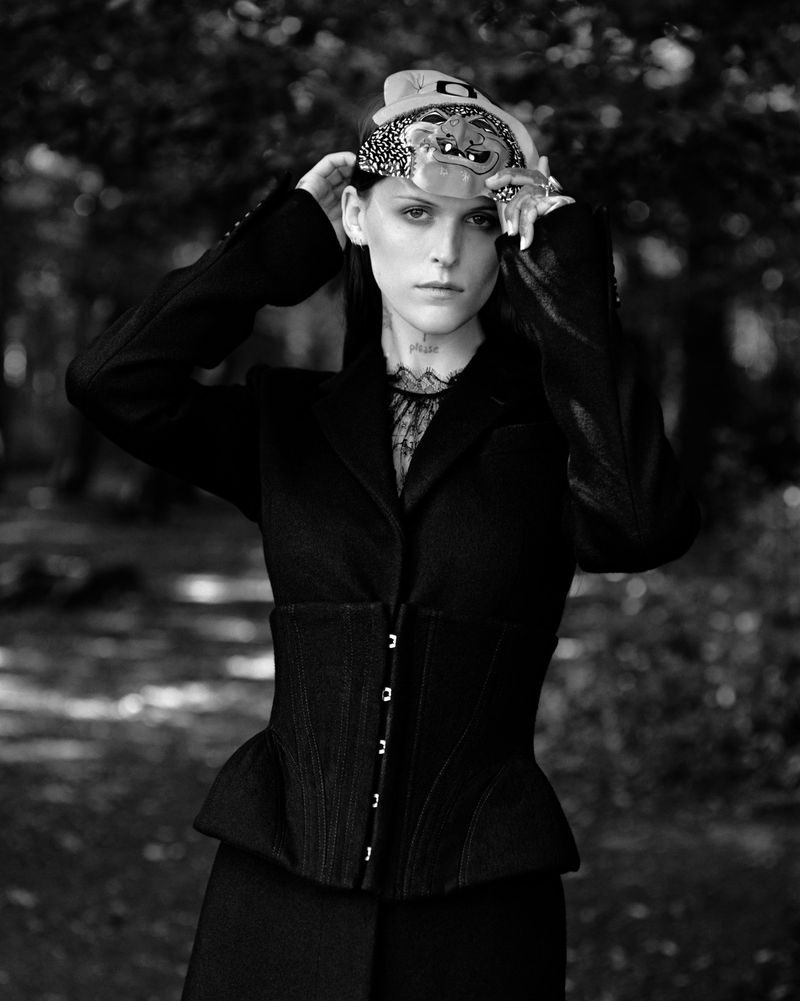 Monochrome portrait of a woman in a black jacket and corset, wearing a decorative headdress, standing in a blurred outdoor setting.