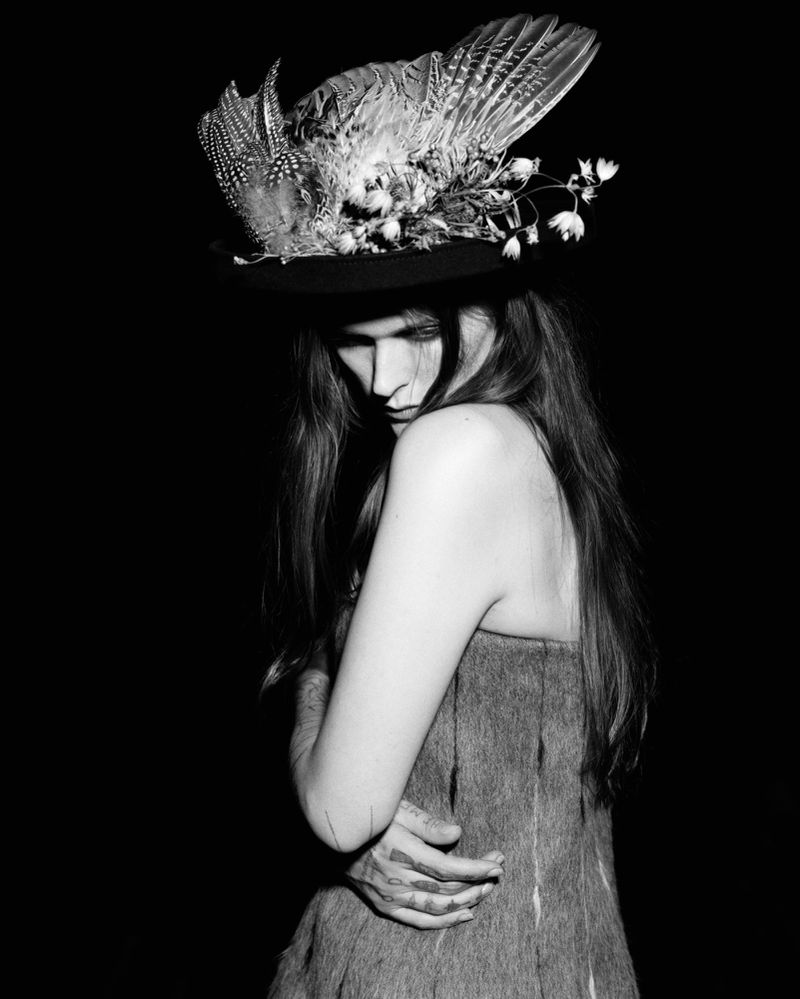 Black and white portrait of a long-haired woman in a fur-like garment and a hat adorned with feathers and flowers against a black backdrop.