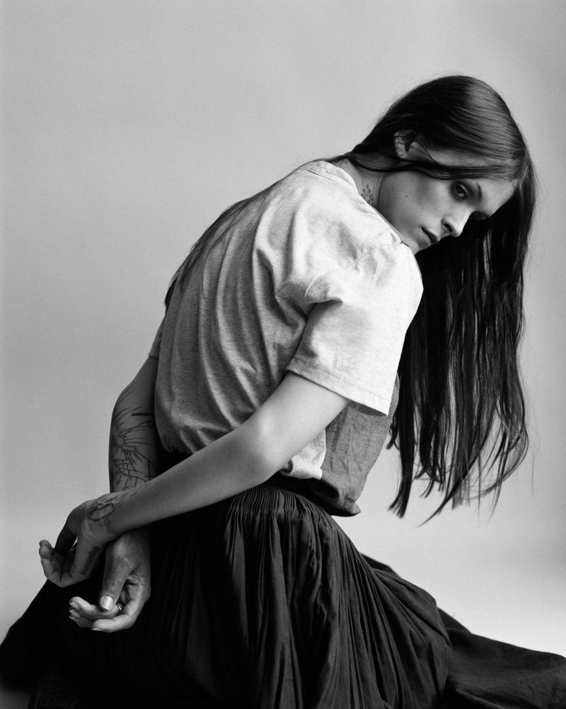 Monochrome portrait of a young adult woman with long hair, looking back over her shoulder while kneeling. She is wearing a t-shirt and skirt.