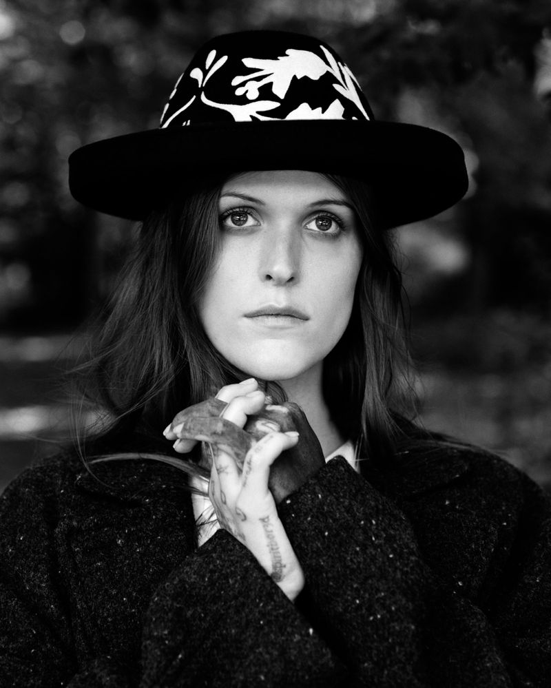 Monochrome portrait of a young woman with long hair wearing a patterned hat and dark coat in an outdoor setting with blurred background.