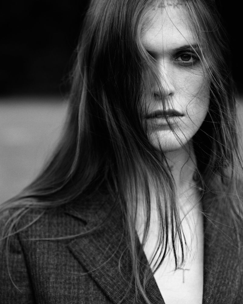 Monochrome portrait of a young adult woman with long hair partially obscuring her face, wearing a textured blazer. Studio lighting.