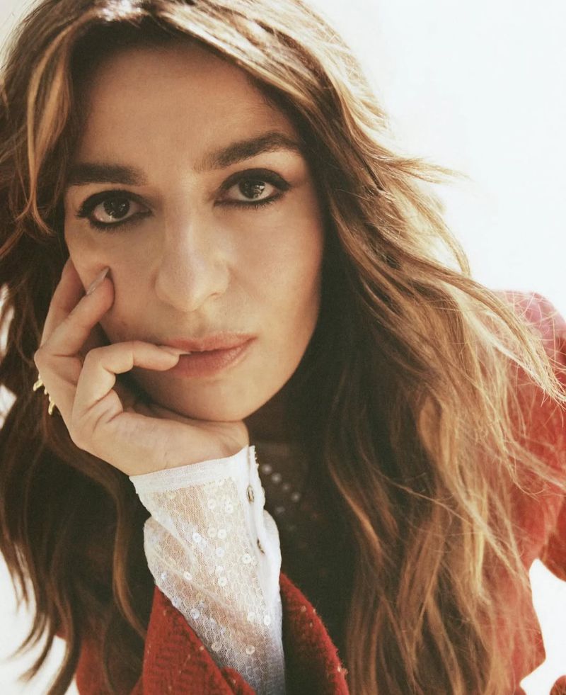 Close-up portrait of an adult woman with light brown hair, wearing a red jacket and white lace top, resting her face on her hand.