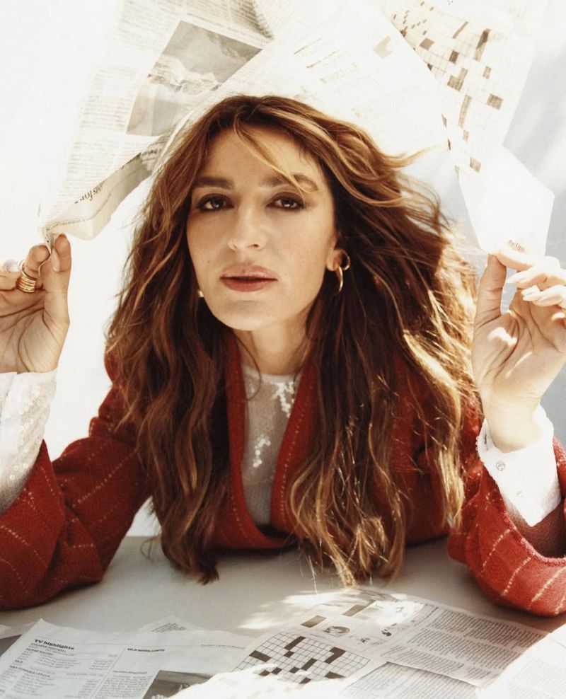 Portrait of a woman with long wavy hair and a red blazer. She is seated at a table with newspapers in the foreground and background.