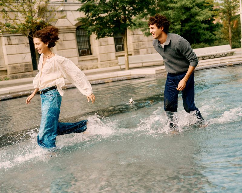 A smiling couple in a light-filled outdoor setting, wading through a shallow pool of water, creating playful splashes on a sunny day.