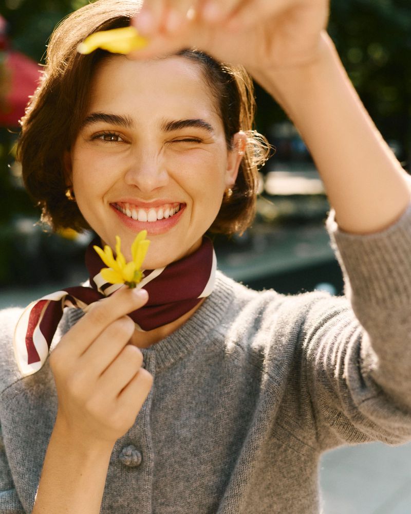 Young woman smiles, holding yellow flower to the camera and winking. She wears a grey sweater, scarf, and gold earrings in an outdoor setting.
