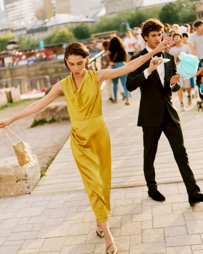 Woman in yellow dress walks beside man in tuxedo holding cotton candy outdoors on cobblestone with a crowd in the background.