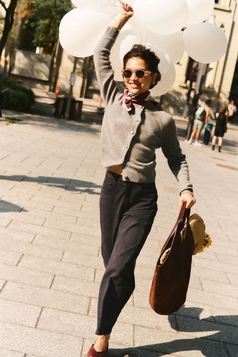 A woman wearing sunglasses and a scarf smiles while walking on a city street, holding a bunch of white balloons and a brown purse.