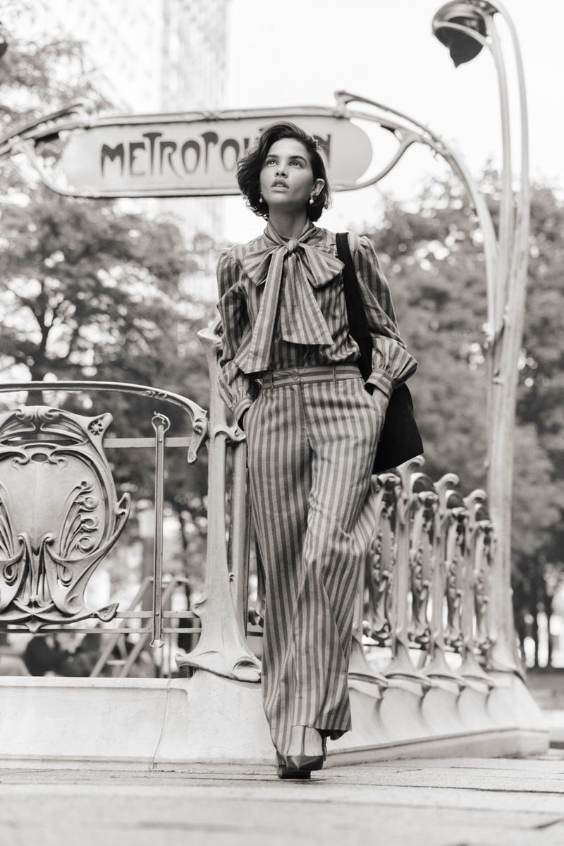 Black-and-white shot of a woman in a striped pantsuit standing in front of a Paris Métro entrance, looking up, with a black tote bag.