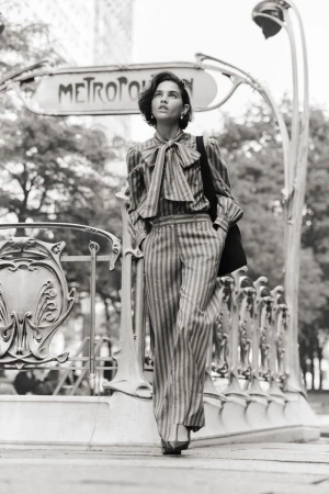 Black-and-white shot of a woman in a striped pantsuit standing in front of a Paris Métro entrance, looking up, with a black tote bag.