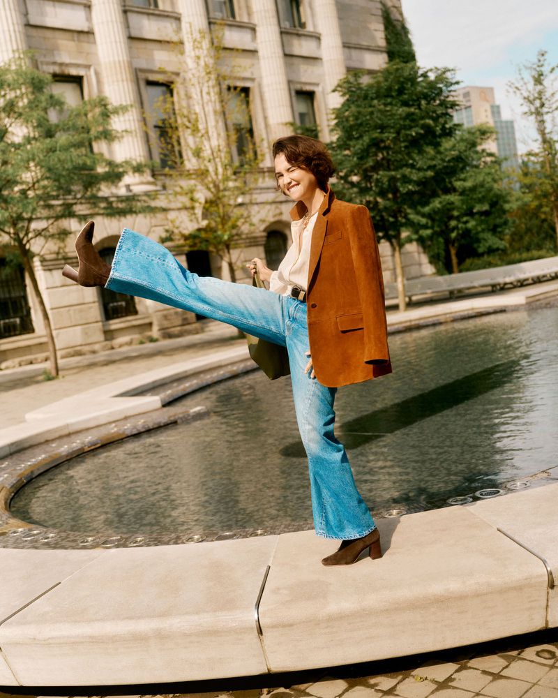 A woman playfully kicks her leg out while balancing on a stone edge, wearing jeans, a brown blazer, and matching boots in a city park.