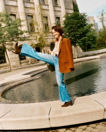 A woman playfully kicks her leg out while balancing on a stone edge, wearing jeans, a brown blazer, and matching boots in a city park.