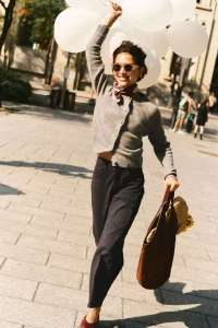 Smiling woman walking outdoors holding white balloons and a brown leather tote bag, wearing a grey cardigan and dark trousers.