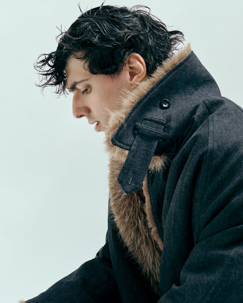 Man in a dark coat with fur collar looking down with wet hair in a photo studio against a bright neutral background.