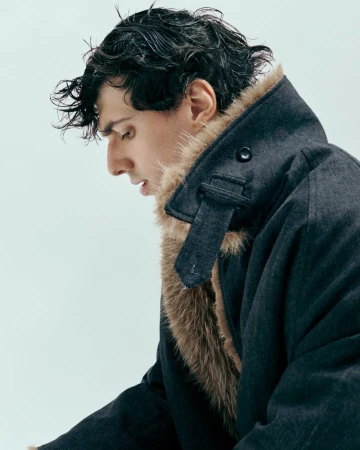 Man in a dark coat with fur collar looking down with wet hair in a photo studio against a bright neutral background.