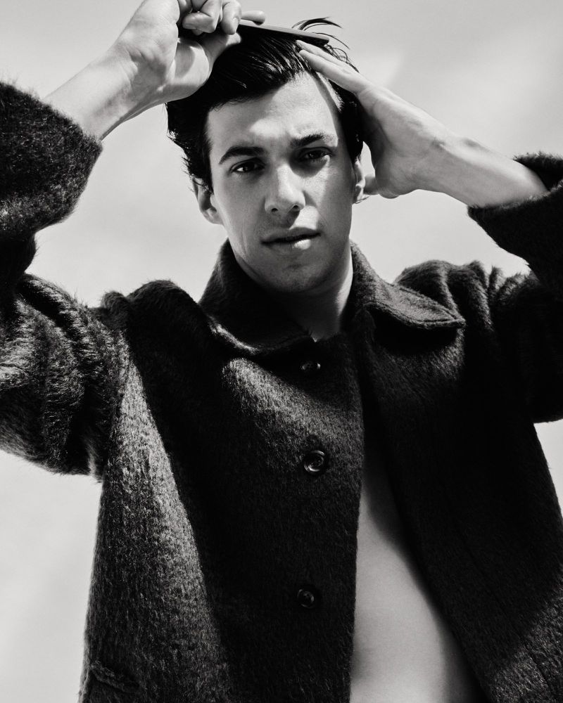 Black and white portrait of a young adult man combing his hair, wearing a dark jacket against a bright sky, showcasing grooming and style.