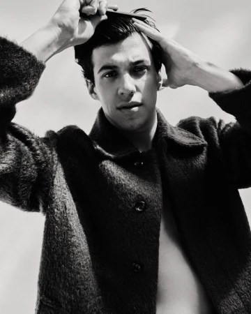 Black and white portrait of a young adult man combing his hair, wearing a dark jacket against a bright sky, showcasing grooming and style.