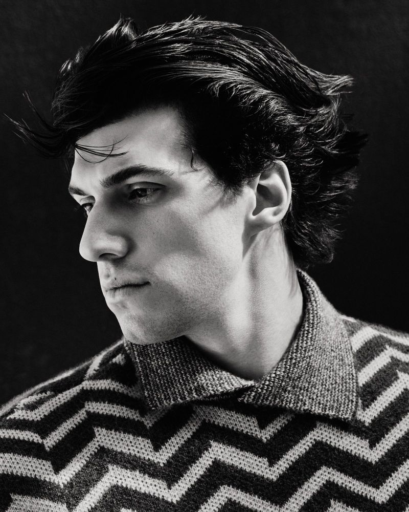 Portrait of a dark-haired man with a patterned sweater looking down and to the left against a dark background in black and white.