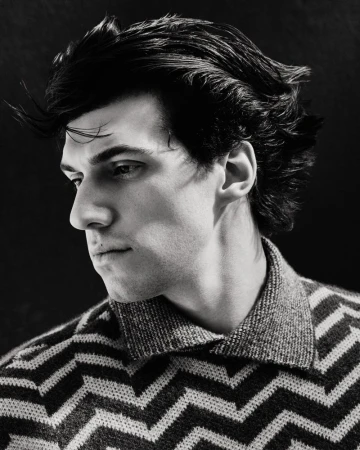 Portrait of a dark-haired man with a patterned sweater looking down and to the left against a dark background in black and white.