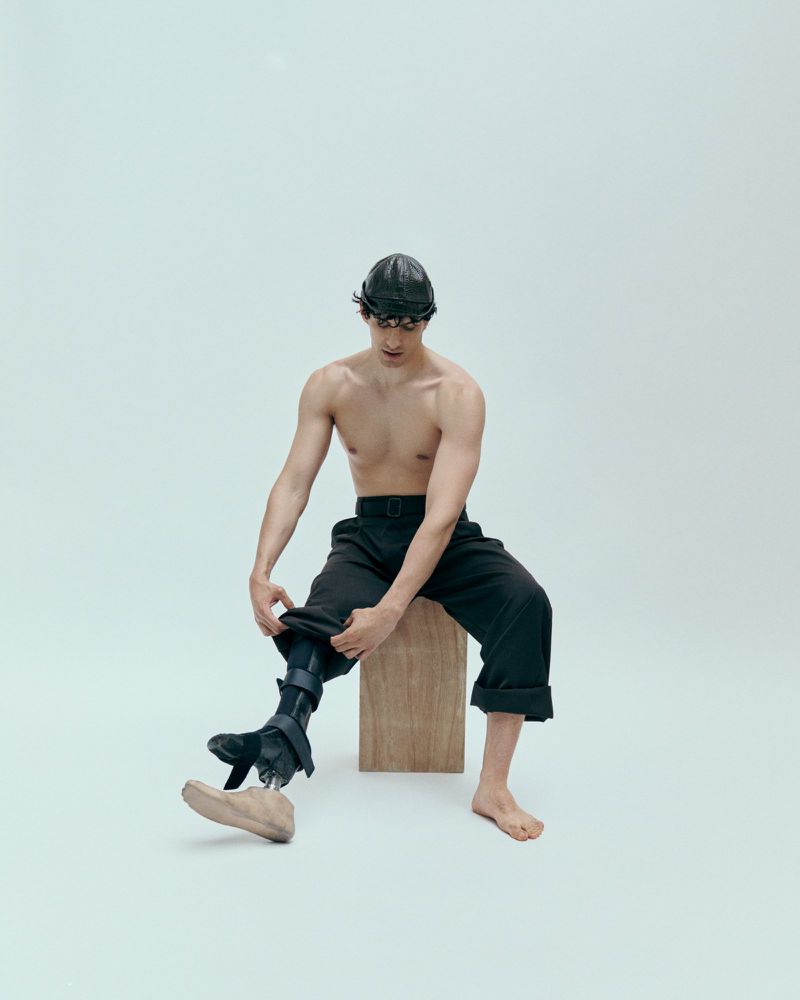 Shirtless man with a prosthetic leg sits on a wooden cube in a bright studio, dressing his prosthetic with a tan shoe.