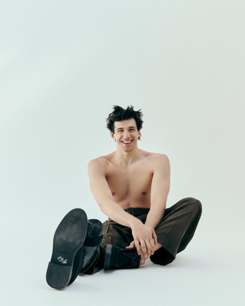 An adult male sits cross-legged on a white background, shirtless and smiling, wearing brown pants and black shoes, looking directly at the camera.