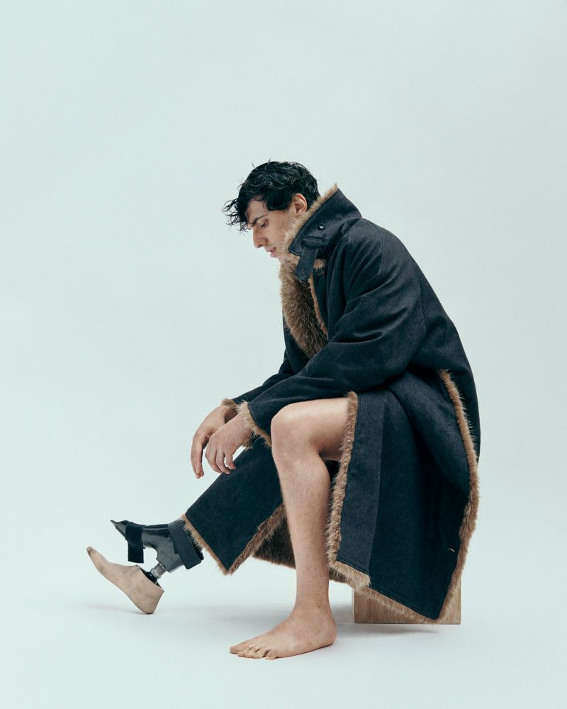 A man with a prosthetic leg poses seated in a studio wearing a fur-lined coat, showcasing fashion and inclusivity in a minimalist style.