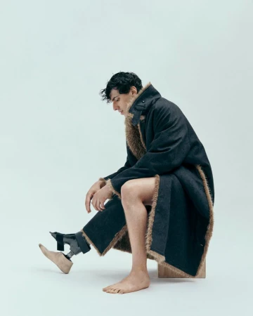 A man with a prosthetic leg poses seated in a studio wearing a fur-lined coat, showcasing fashion and inclusivity in a minimalist style.
