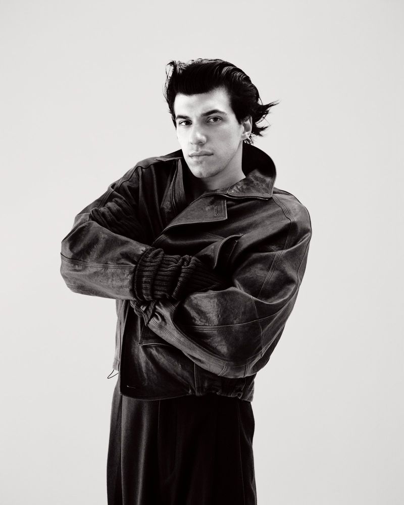 Monochrome studio portrait of a young adult man with dark hair in a leather jacket, posing with crossed arms against a light background.
