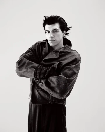 Monochrome studio portrait of a young adult man with dark hair in a leather jacket, posing with crossed arms against a light background.