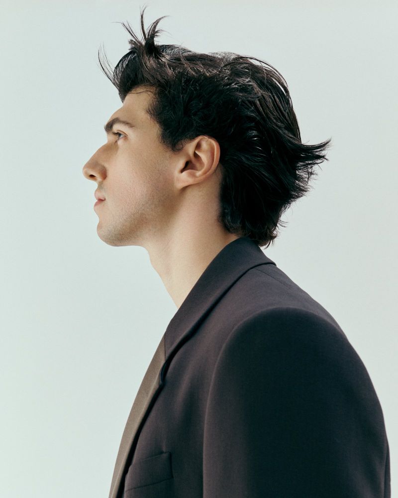 Side view portrait of a young adult man with dark hair wearing a dark blazer against a light blue backdrop in a studio setting.