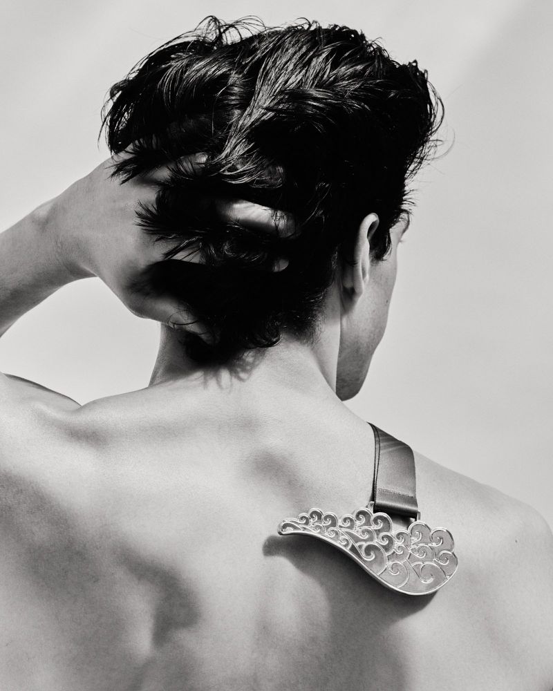 Monochromatic back shot of a shirtless adult male model with dark hair, wearing a decorative silver pendant on a dark strap.