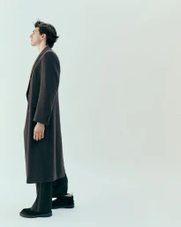 Man wearing a long dark coat and black trousers standing against a plain white seamless studio background.