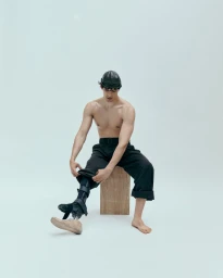 Shirtless man wearing dark trousers and cap sitting on a wooden stool against a white seamless background