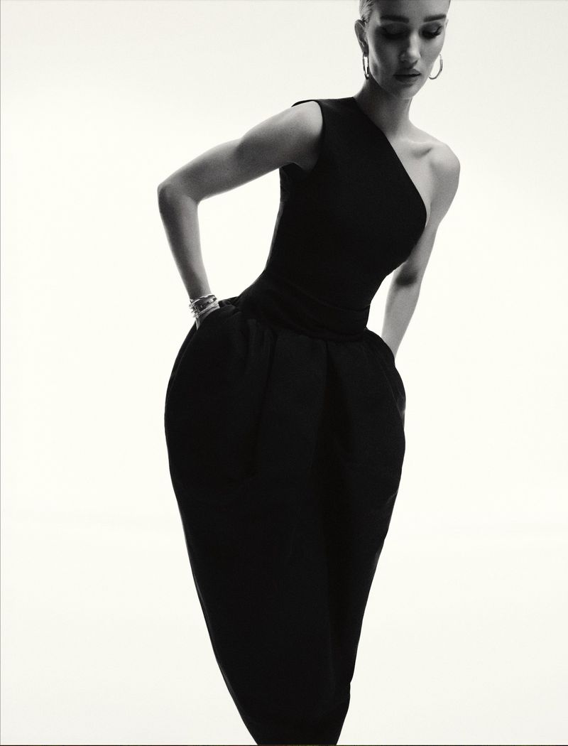 A stylish adult woman in a black one-shoulder dress poses against a bright white backdrop in a fashion portrait with an elegant look.