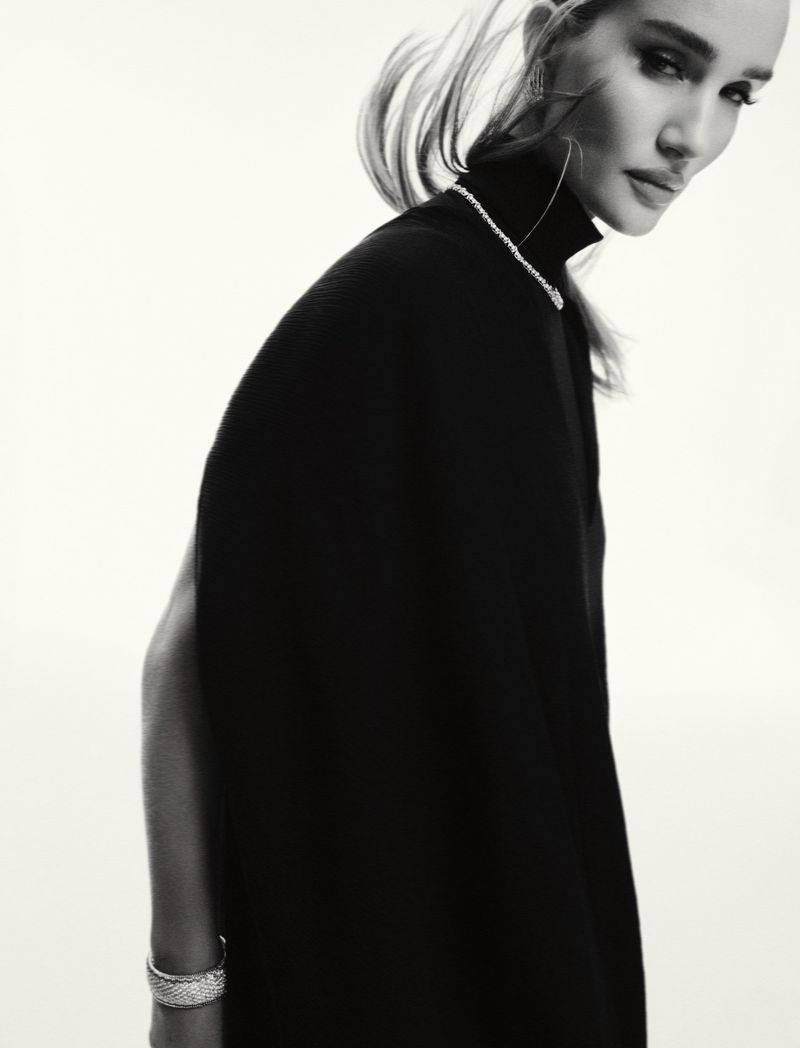 Black and white portrait of a woman with blonde hair wearing a dark turtleneck top and jewelry against a bright neutral background.