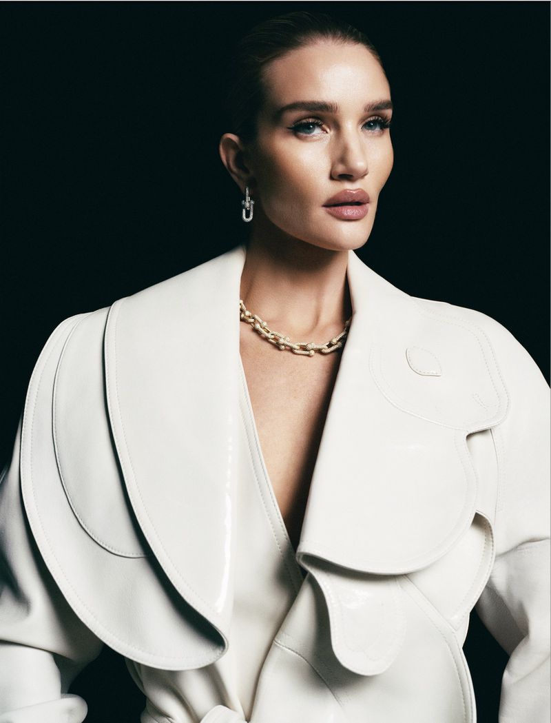 Rosie Huntington-Whiteley in a cream trench coat, gold chain necklace, and drop earrings against a dark backdrop. Beauty and fashion shot.