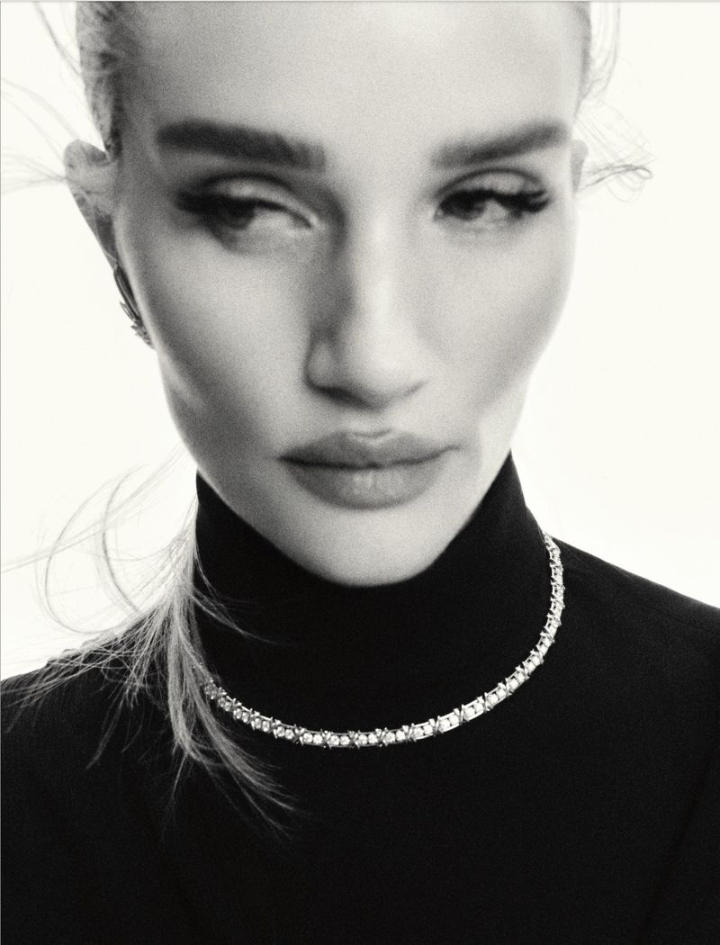 Close-up studio portrait of a woman with elegant makeup, wearing a black turtleneck and a crystal necklace against a bright background.