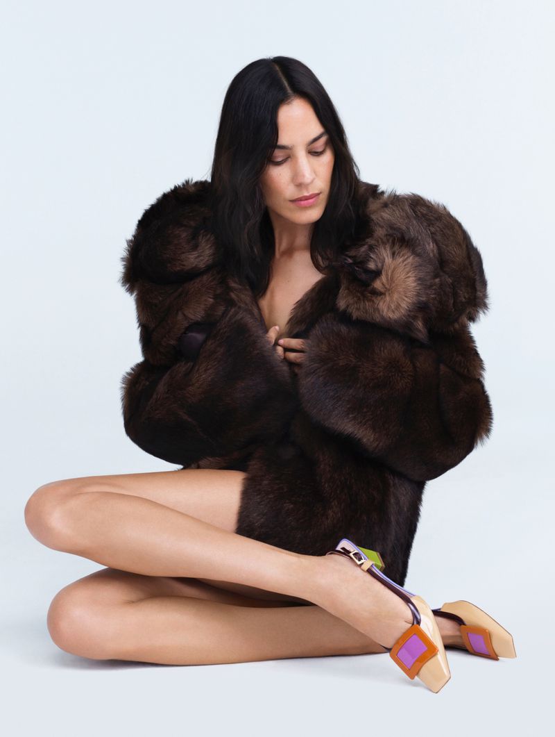 A woman with dark hair sits in a fur coat and colorful heels in a studio setting, exuding fashion and style against a white backdrop.