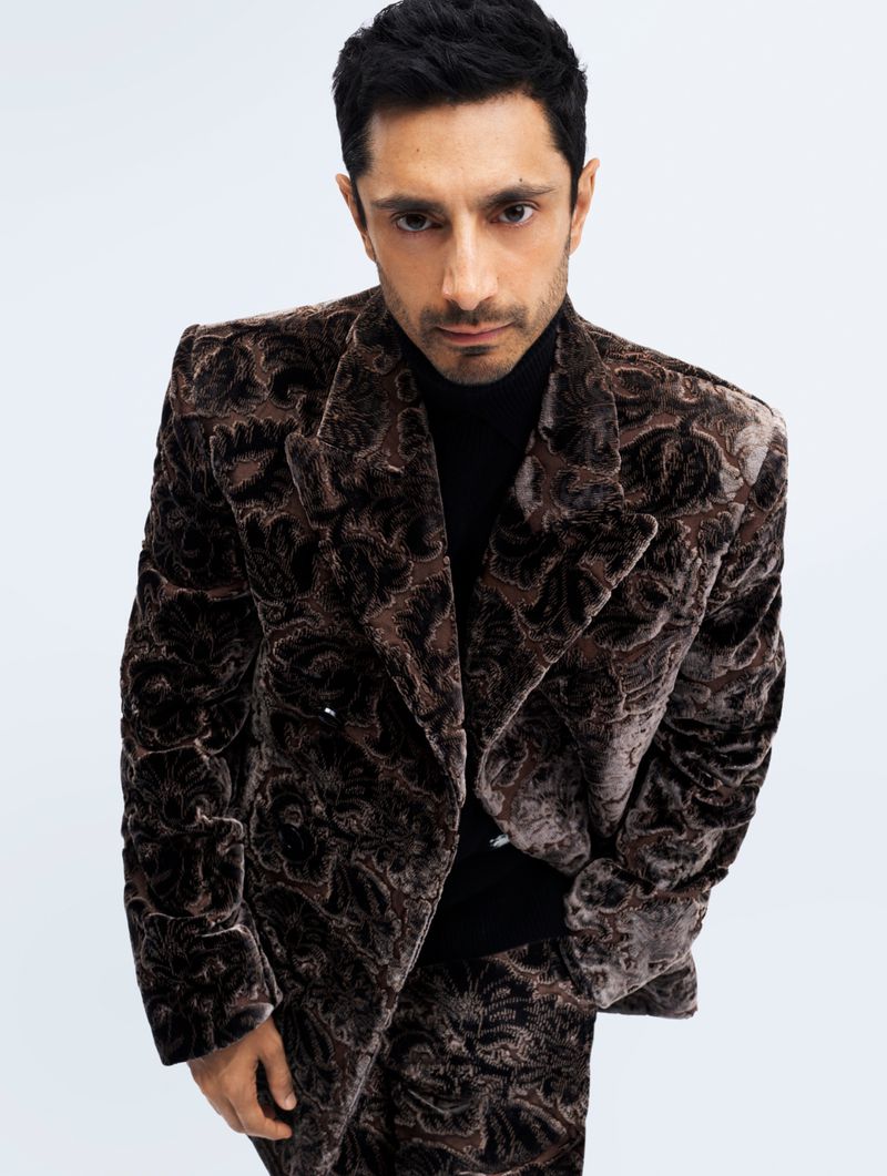 Portrait of a man in a patterned brown suit and black turtleneck against a white backdrop, looking directly at the camera.