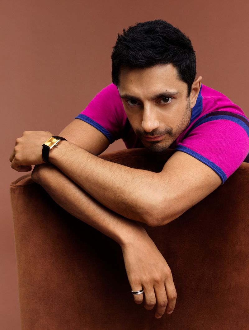 Close-up portrait of a man with dark hair leaning on a brown velvet chair, wearing a pink and blue shirt and a gold watch.