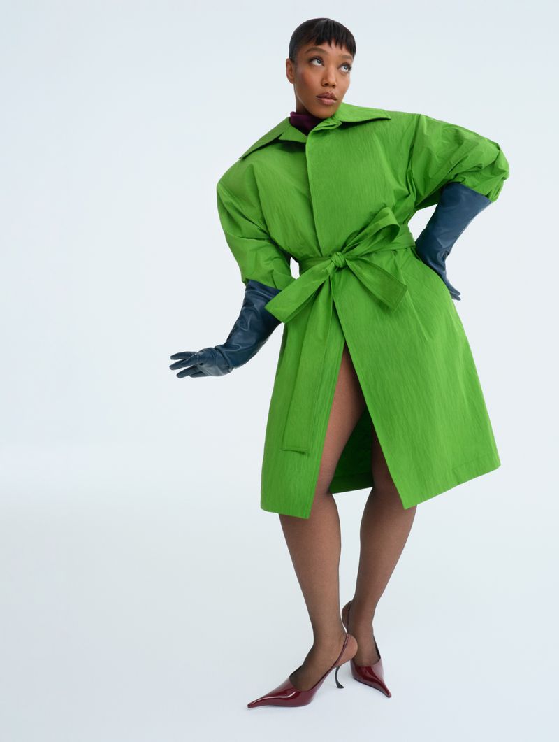 An adult woman in a vibrant green coat, gloves and matching pumps, posing against a clean white backdrop in a fashion-forward image.
