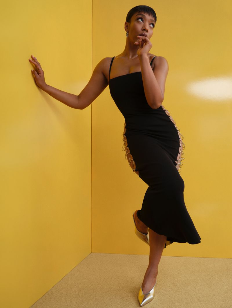Elegant woman in black dress and gold heels strikes a pose against a vibrant yellow wall, blending fashion with modern art.