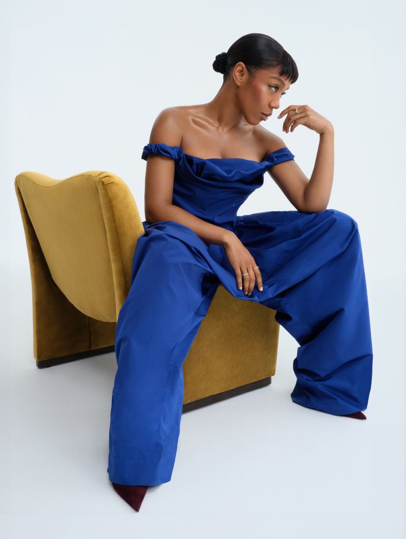 Woman sits on yellow chair wearing blue off-the-shoulder jumpsuit in a photo studio with soft, diffused lighting and a minimalist aesthetic.